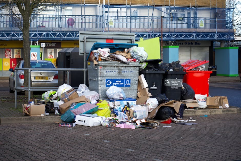 A large pile of mixed household waste and recyclable materials is scattered around a series of trash bins on a paved urban street. The waste includes cardboard boxes, plastic bags, paper, and various packaging materials, some spilling onto the nearby sidewalk. The bins consist of a grey bin labeled for paper and cardboard, a black bin for general waste, a red bin, and an additional black bin, all positioned on the curbside. Behind the waste scene, a section of a retail or commercial building with storefronts and signage is visible, along with a parked silver car situated behind a metal railing. An adjacent tree is partially seen on the left side of the image, with branches extending upward. The area appears to be an outside designated rubbish collection point, illustrating a typical scenario for independent waste disposal or rubbish removal services, such as those provided by rubbishclearanceswisscottage.com, emphasizing the importance of proper waste handling and collection.