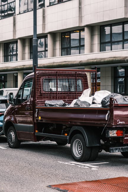 A small burgundy flatbed truck parked on a city street in front of a modern multi-storey office building with large rectangular windows. The truck's bed is loaded with various types of waste, including several large black and white plastic bags, some of which are partially opened revealing contents. The waste appears to consist of household or construction debris, and the textured plastic bags contrast with the smooth, metallic finish of the truck. The truck features a protective metal grid at the cab partition, and its rear lights and wheel arches are clearly visible. The surrounding environment includes a street lamp and partial views of other vehicles, with a clear, daytime setting. This scene illustrates an example of off-site, private rubbish collection, highlighting alternative waste handling options often arranged through rubbish clearance services like those potentially offered by companies operating in Swiss Cottage.
