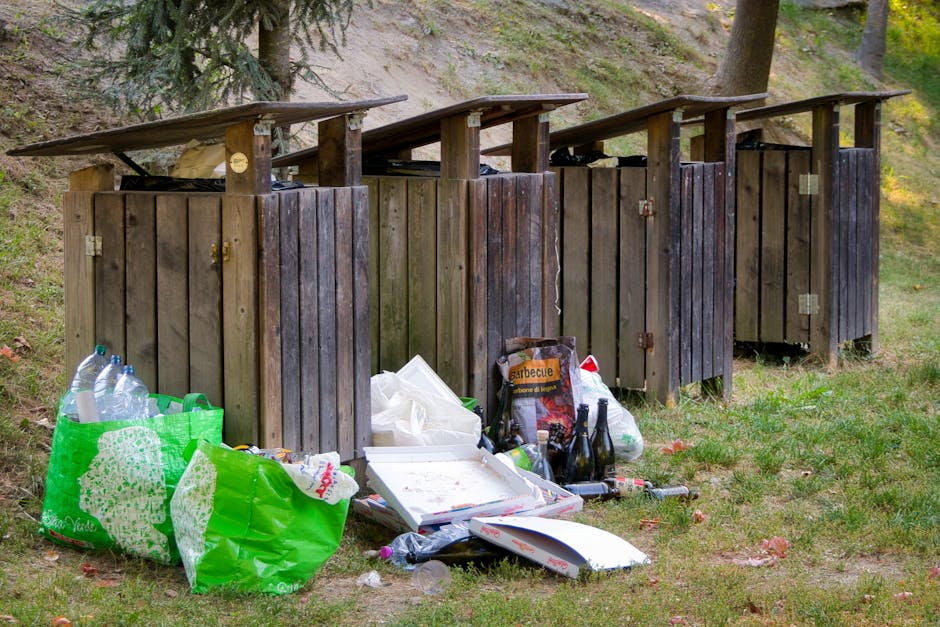 A row of four small wooden outdoor trash enclosures with sloped roofs, situated on a grassy area with sparse fallen leaves and surrounded by trees. The enclosures are constructed from vertical wooden planks stained in dark brown, with hinges and locks visible on some doors. In front of the enclosures, there are various discarded items including three green plastic bags filled with empty plastic bottles, a white cardboard pizza box with food remnants, a black plastic bag, and several glass bottles, some dark and some clear. The background shows a hillside with patches of grass and a few mature trees, with sunlight filtering through the foliage, creating a natural outdoor scene that highlights the presence of rubbish left outside the designated storage units, reflecting a context of informal waste disposal or improper rubbish handling.