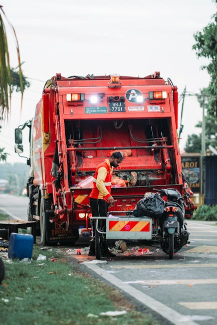 A sanitation worker dressed in a yellow and red high-visibility safety vest operates a large red waste collection truck parked on the side of a roadway, during daytime. The worker is using a tool to load or unload waste, with a black plastic bag placed on a small metal platform attached to the truck. The scene includes some scattered debris on the pavement and nearby, as well as a motorbike parked close to the waste collection area. The background features trees, a road with passing vehicles, and power lines, indicating an urban or suburban environment. The lighting appears natural, with an overcast sky providing diffuse illumination. This setting illustrates a private waste collection operation, potentially an alternative to local authority rubbish disposal, with the truck prominently branded and the worker actively handling waste materials consistent with rubbish clearance services.